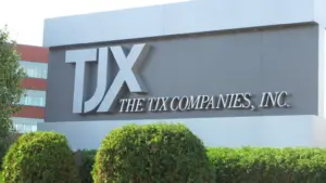 Outdoor sign for The TJX Companies, Inc. with large silver letters on a grey background, framed by green bushes.