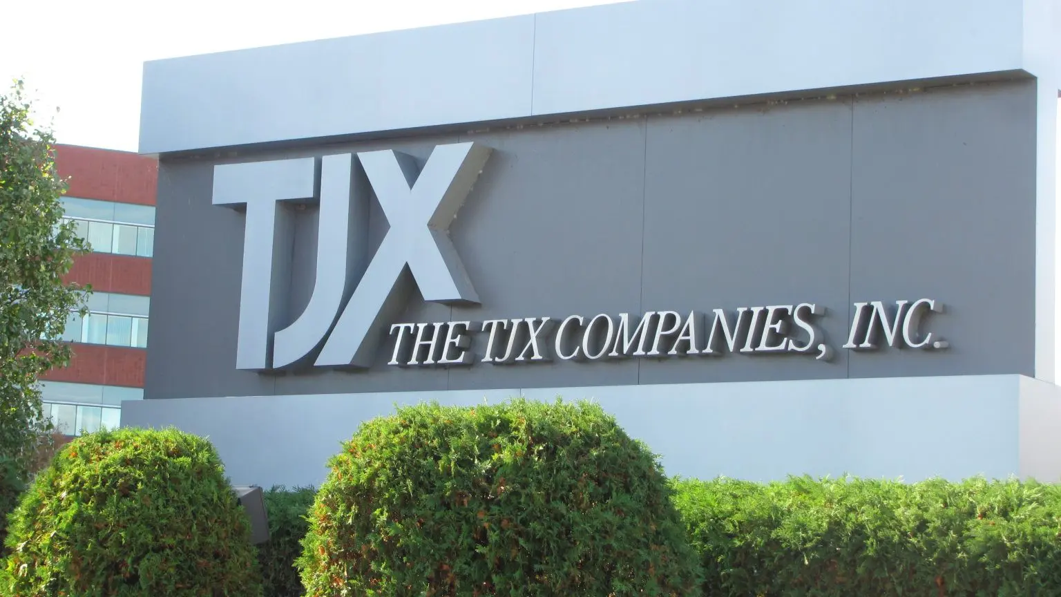 Outdoor sign for The TJX Companies, Inc. with large silver letters on a grey background, framed by green bushes.
